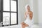 A beauty-loving woman wearing a towel with a healthy, smooth body looking at a round mirror in a white bathtub in a bathroom