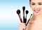 Beauty portrait of lovely beautiful happily smiling woman holding a bunch of make-up brushes against a light blue background