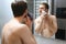 beauty, shaving, grooming and people concept - close up of young man looking to mirror and shaving beard with trimmer or