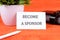 BECOME A SPONSOR text written on a business card next to a business card holder, a green flower and a pencil on an orange