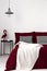 Bed dressed in burgundy organic cotton bedding and vanilla soft blanket in a modern bedroom interior with white empty wall. Real