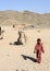 The bedouin girl walking alone in the desert against the background of a lying camel and hills.