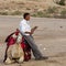 A bedouin tourist guide is leaning onto his camel waiting for tourist customers.