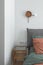 Bedroom interior with rust color linen and cushions on a bed, grey blanket, rattan bedside table and metal wall light.