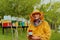 The beekeeper holding a jar of honey in his hand while standing in a meadow surrounded by a box and a honey farm