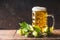 Beer mug with cap of foam on table with fresh hops at dark rustic background