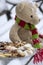 Beige plushy teddy bear with red green striped knitted scarf sitting with Christmas cookies on the bench covered with white snow