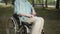 Being in the public park, a disabled white man in a blue shirt and a protective mask is sitting a wheelchair with a