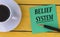 BELIEF SYSTEM - word on a green sheet on a yellow wooden background with a pen and a cup of coffee