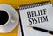 BELIEF SYSTEM - words on a white sheet on a yellow wooden background with a pen and a cup of coffee