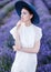 A belted portrait of a beautiful girl in a white dress and a blue hat standing in the middle of blossoming lavender fields and