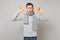 Bemused young man in gray sweater, scarf holding oranges on grey wall background, studio portrait. Healthy