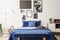 Bench in front of bed with navy blue pillows between lamp and cabinet in bedroom interior. Real photo