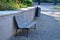 A bench with a gray paneled seat by the retaining wall on a granite cube sidewalk in a city park and a metal trash can