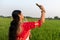 A Bengali girl wearing a red and white sari is taking a selfie by her cell phone in a vast paddy field