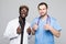 Best doctors. Two caucasian and afro american smiling doctors show a thumbs up while standing against gray background