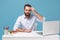 Bewildered young bearded man in light shirt sit and work at desk with pc laptop isolated on pastel blue background