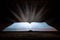 The big book of the Bible lies on a wooden table. In the dark. A light shines on the book from above. Light comes out of the book