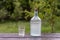 Big bottle and glass of moonshine or vodka on wooden table in sunny summer day in garden, close up