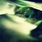 Big boulders covered by fresh green moss in foamy water of mountain river. Light blurred cold water with reflections, white