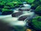 Big boulders covered by fresh green moss in foamy water of mountain river. Light blurred cold water with reflections, white