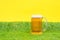 Big mug of beer standing on green grass of artificial lawn with bright yellow background. Copy space. Oktoberfest