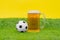 Big mug filled with cold beer with foam, and miniature soccer ball stand on grass of artificial lawn on yellow