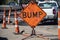 Big orange grungy sign that says BUMP surrounded by highway construction cones with trucks and torn up pavement in the background