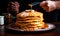 Big pile of freshly fried thick pancakes with caramel sauce. Hand of a cook holding a spoon over the breakfast. Close up.