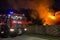 Big red fire truck parked at brick fence on burning wooden barn at night. High orange fire flames, dense smoke on dark sky, trees