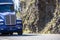 Big rig blue powerful semi truck running on the winding road with rock wall