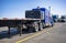 Big rig blue semi truck with empty flat bed semi trailer standing on truck stop parking lot opposite another semi trucks standing