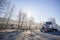 Big rig classic American semi truck with covered dry van semi trailer going on the winter road with frosted trees in sunshine