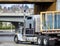 Big rig classic semi truck transporting glass sheets in wood boxes on flat bed semi trailer turning under the bridge