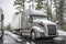 Big rig gray semi truck with dry van semi trailer standing on the wet winter truck stop parking lot with snow and ice on the cab