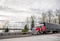 Big rig red classic bonnet semi truck transporting cargo in covered rubberized black semi trailer driving on the road intersection