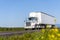 Big rig white day cab semi truck for local deliveries with long box semi trailer driving on the summer road with yellow flowers