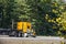 Big rig yellow semi truck with step down semi trailer running on the highway with green and autumn trees