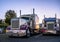 Big rigs classic bonnet semi trucks standing in row on truck stop parking lot at evening time reflecting sunset and all around by