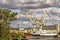 Big ship with rusty sides moored at the pier. Large port cranes form the letter W against a blue sky with heavy dark clouds