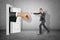 Big stretched fist appearing through white doorway and businessman making kicking gesture on grey wall background