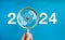 The big white 2024 year number with target dart icon inside the golden magnifying glass in hand on blue background.