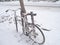 Bike parked next to the tree in the winter. Snow falls on the bike and the road surface becomes white