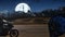 Biker riding the motorcycle at night on the road and the moon is glowing behind the mountain on the dark sky.