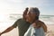 Biracial senior man with arm around woman standing by bicycle looking away at beach on sunny day