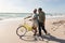 Biracial senior man walking and talking with woman wheeling bicycle at sunny beach against sky