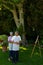 Biracial senior wife tying husband\\\'s apron standing by canvas and easel against plants in yard