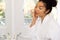 Biracial woman in bathrobe looking in mirror in sunny bathroom