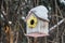 A birdhouse for birds from white and yellow and pink boards with white snow on the roof is on a brown tree in a park in winter