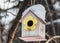 A birdhouse for birds from white and yellow and pink boards with white snow on the roof is on a brown tree in a park in winter